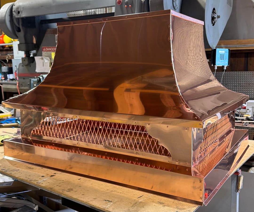 Polished copper kitchen range hood displayed on a wooden workbench.