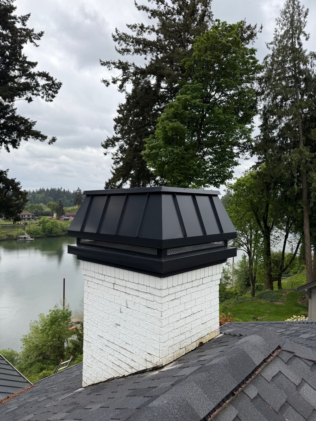Modern black chimney cap on a white brick chimney overlooking a lake and trees.
