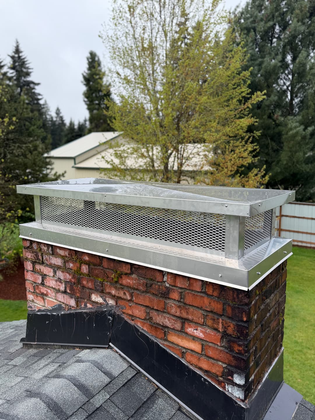 Chimney cap made of stainless steel on a brick chimney surrounded by trees and a grassy yard.