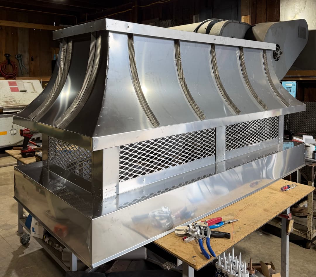 Stainless steel commercial kitchen hood on a workbench in a workshop setting.