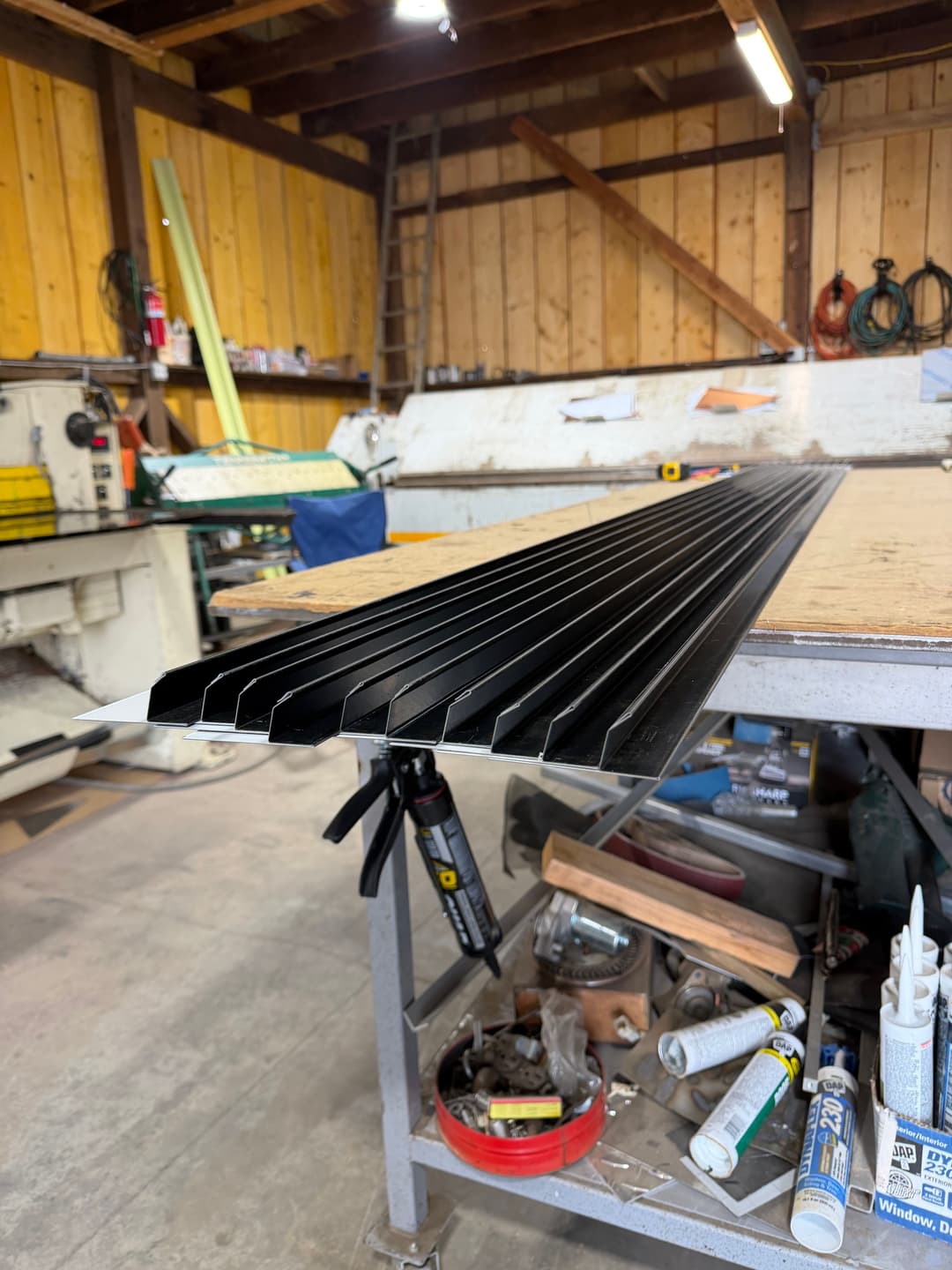 Black metal sheets on a workshop table with tools and equipment in a wood-paneled space.