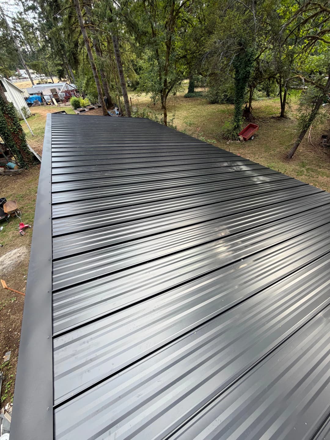 Aerial view of a new metal roof in a wooded area, showcasing sleek black panels and surrounding landscape.