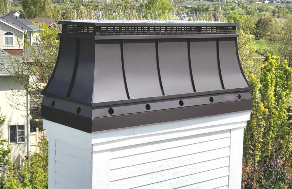 Modern metal chimney cap on a white chimney against a green landscape backdrop.