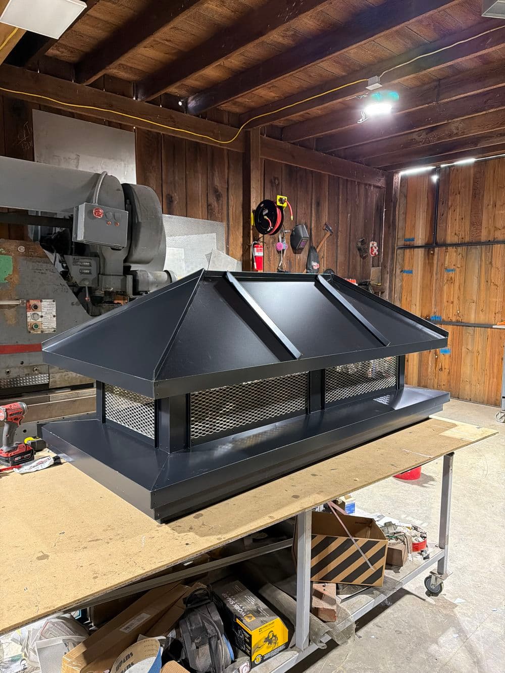 Black steel chimney cap with mesh, displayed on a workbench in a wooden workshop.