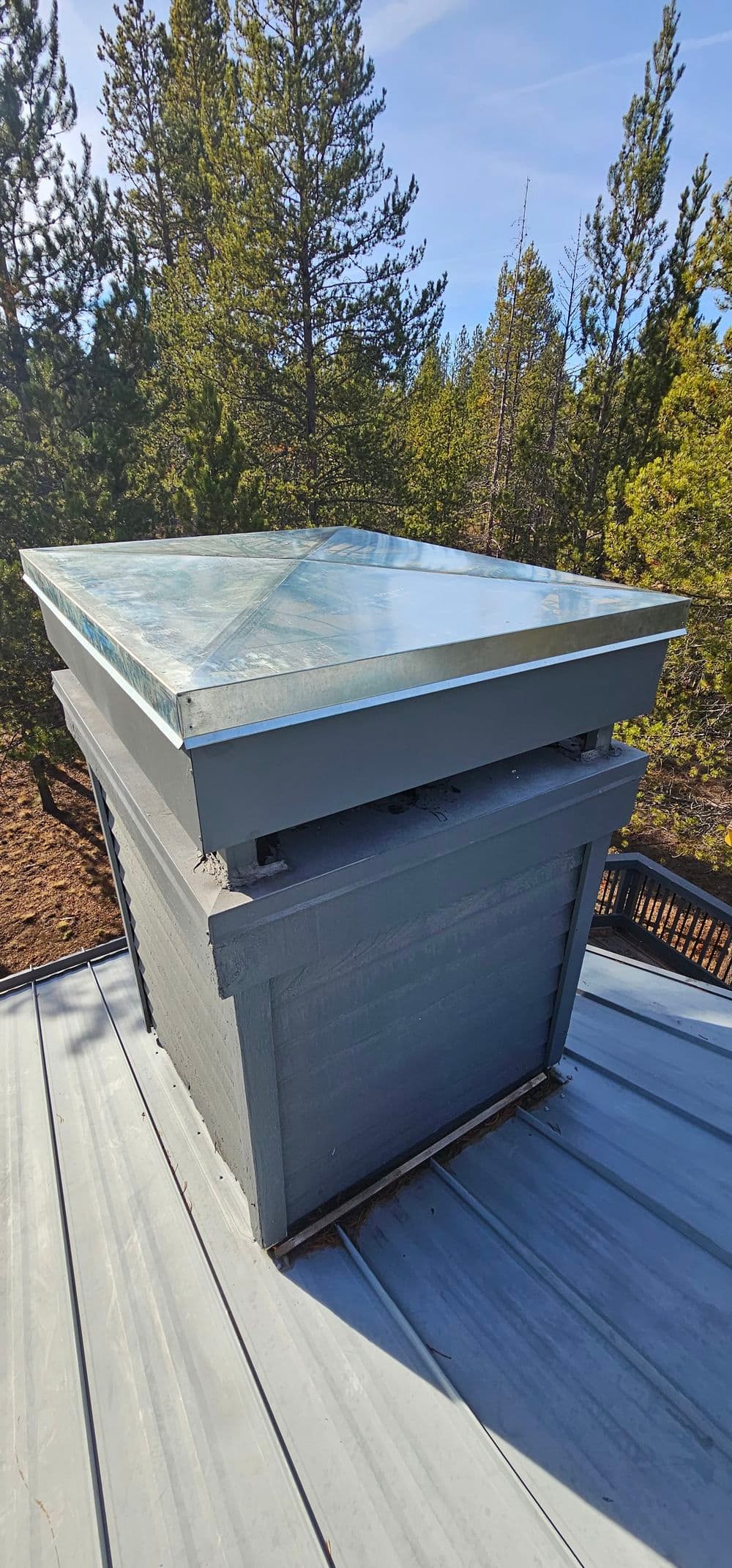 Chimney cap on a rooftop with forest backdrop under clear blue sky.