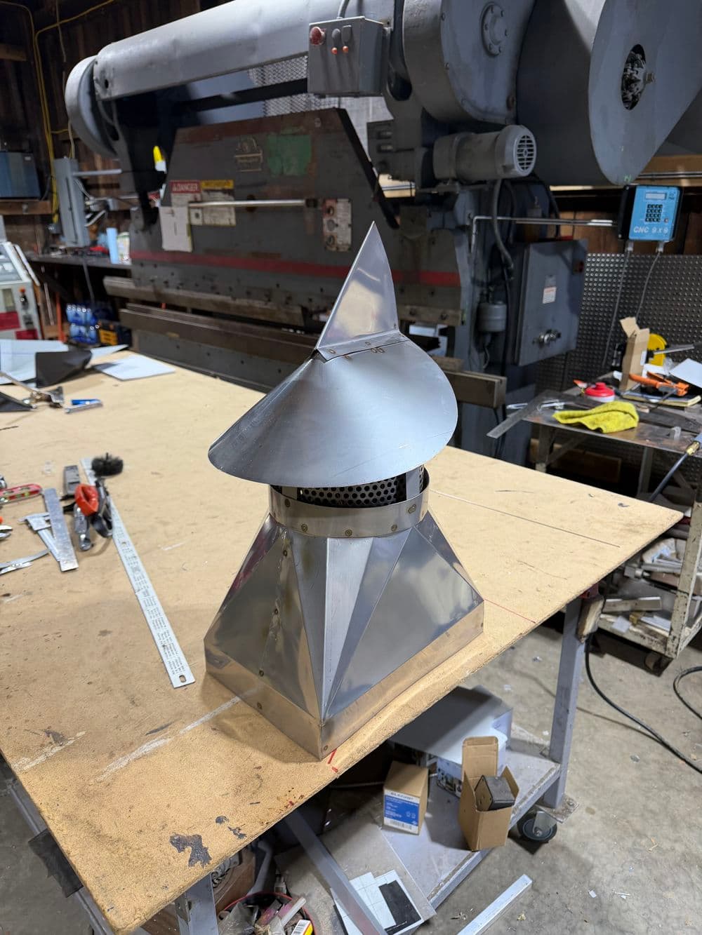 Metal chimney cap on a workbench with machinery in the background, showcasing craftsmanship.