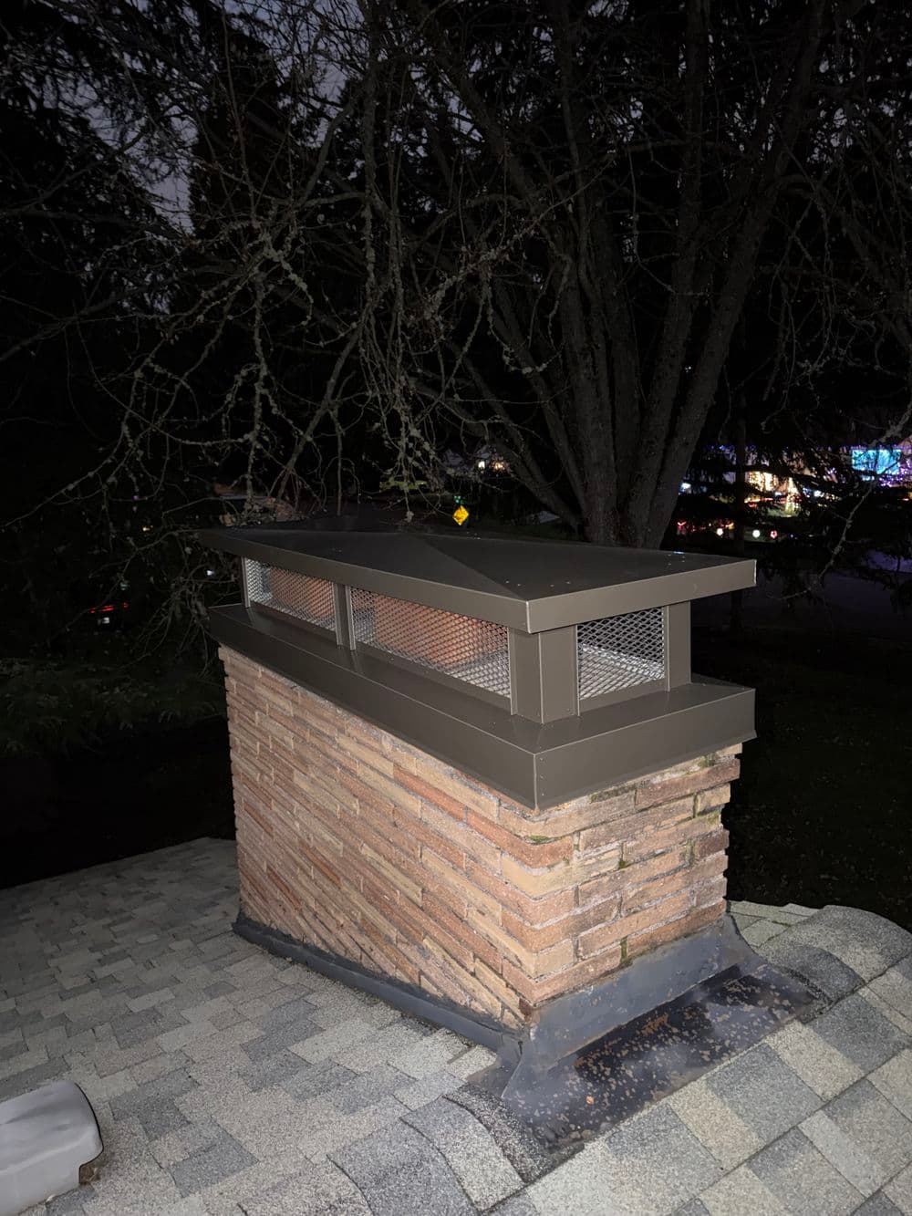 Chimney cap at night with brick structure and surrounding trees, enhancing home ventilation.
