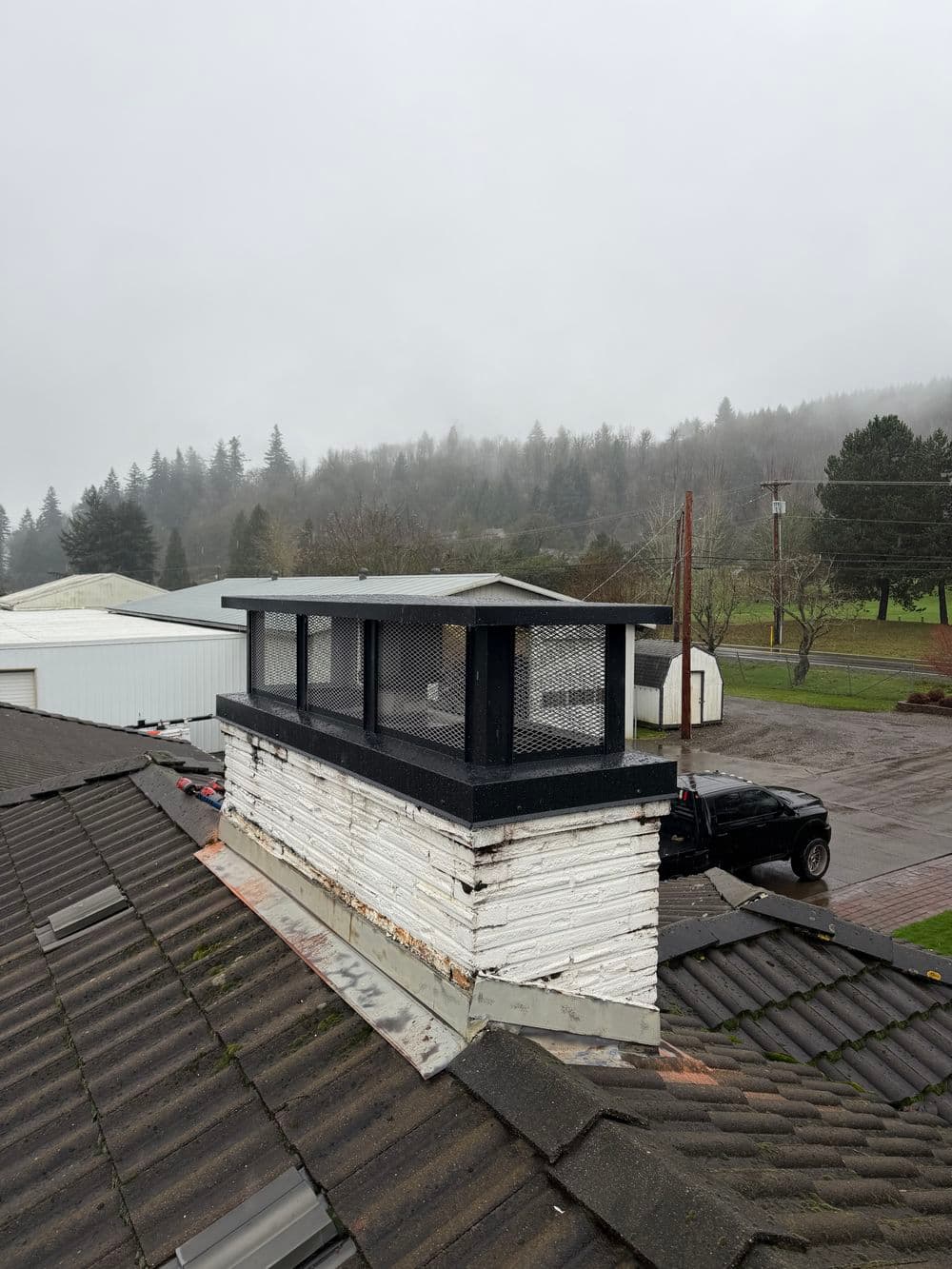 Chimney repair with metal cap on a foggy roof, overlooking tree-dense background.