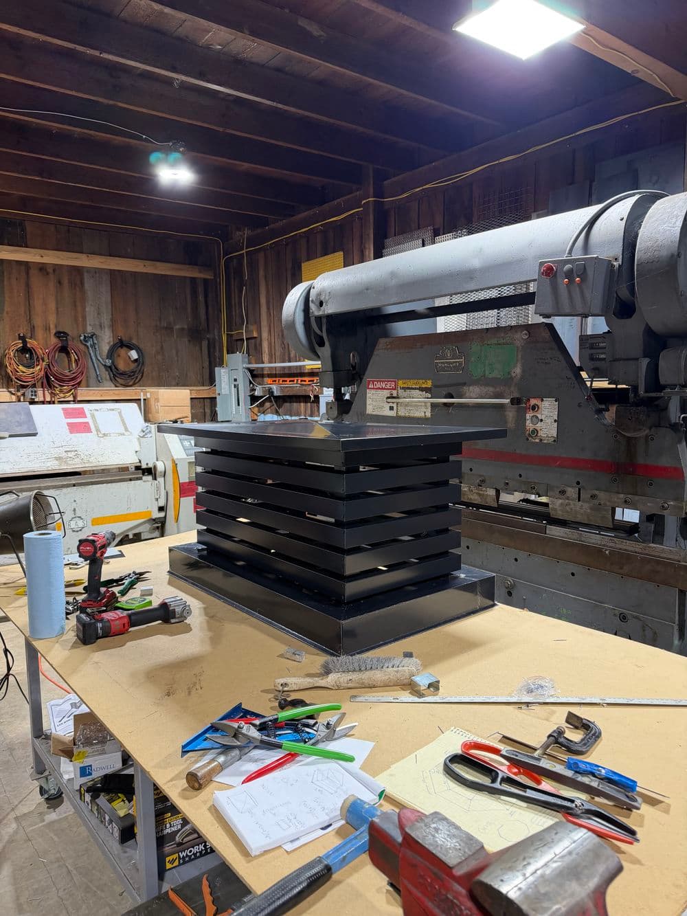 Industrial workshop with a stack of metal sheets on a table, tools, and machinery in the background.